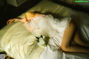 A woman in a white dress sitting on a bed holding a flower.