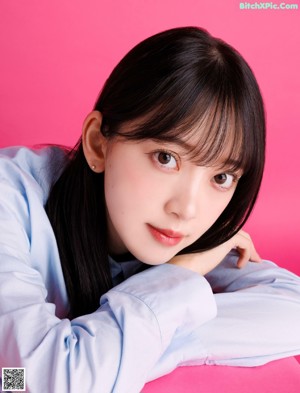 A woman in a blue shirt and gray skirt sitting on a pink background.