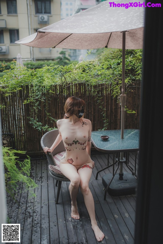 A naked woman sitting on a chair under an umbrella.